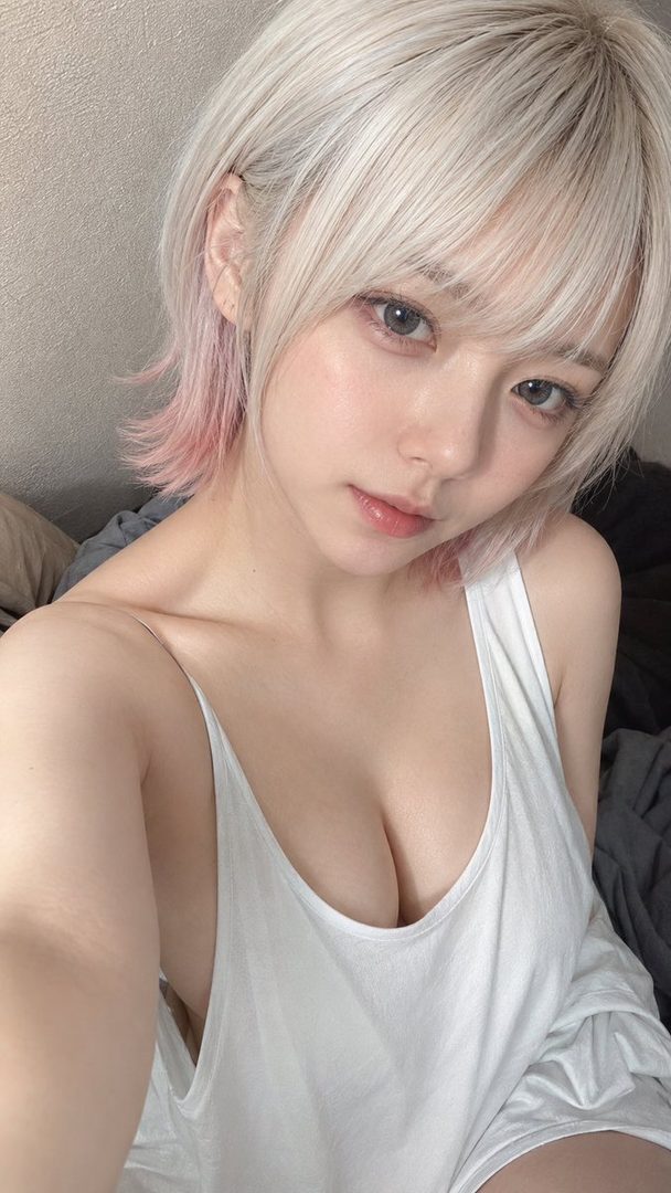 A photorealistic selfie of a young woman with short blonde hair and pink highlights, wearing a white tank top, looking directly at the camera.
