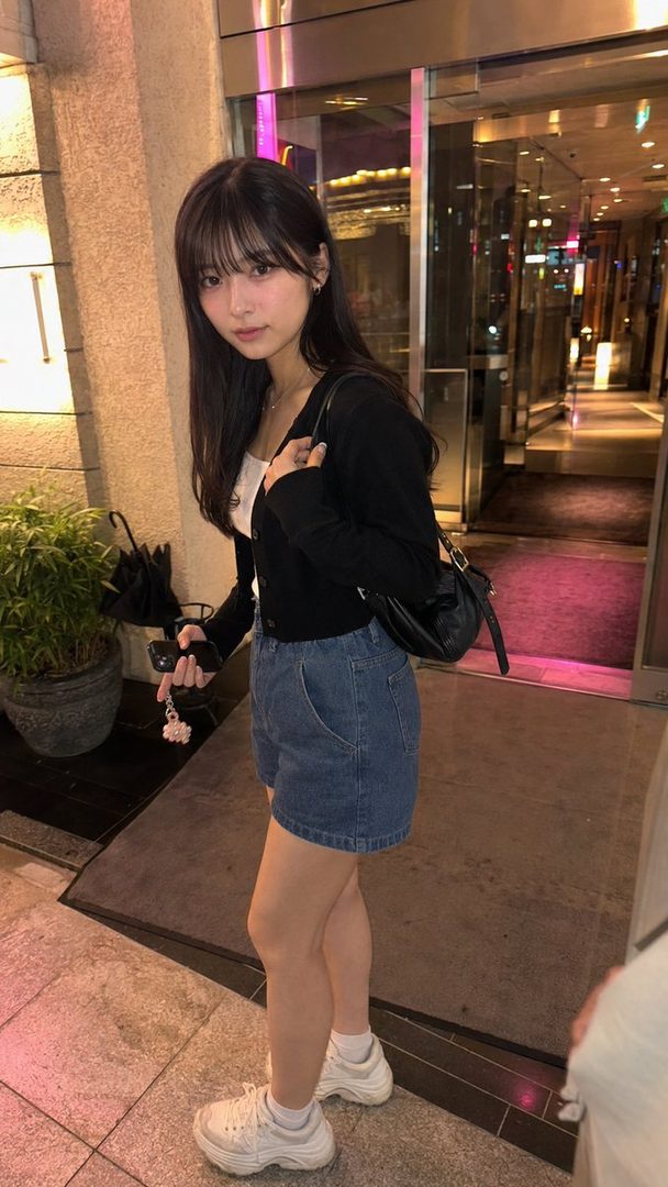 A young woman with long black hair and bangs, wearing a black cardigan, white top, and denim shorts, stands on a city street at night.