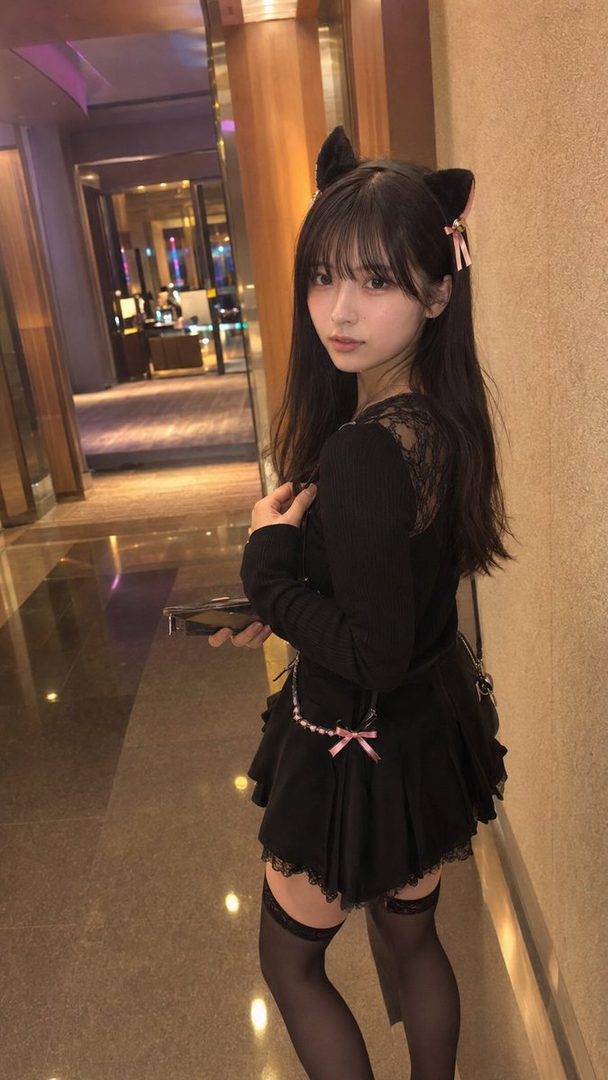 A young woman with long black hair and cat ears poses indoors, wearing a black top, short skirt, and thigh-high stockings.