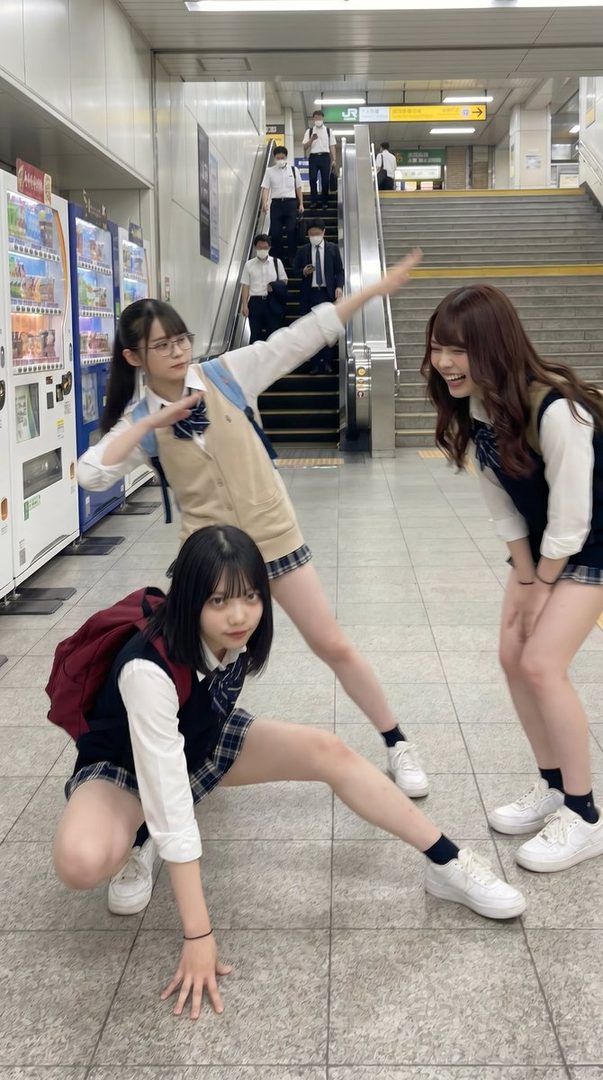 Three young women in Japanese school uniforms pose playfully in a train station, with one in a squatting pose and another with arms outstretched.