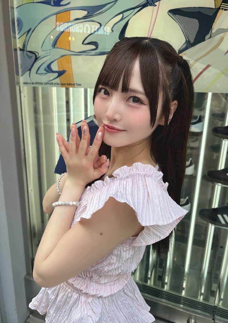 A cute Japanese woman with brown hair in pigtails and bangs, wearing a light pink ruffled top, poses indoors.