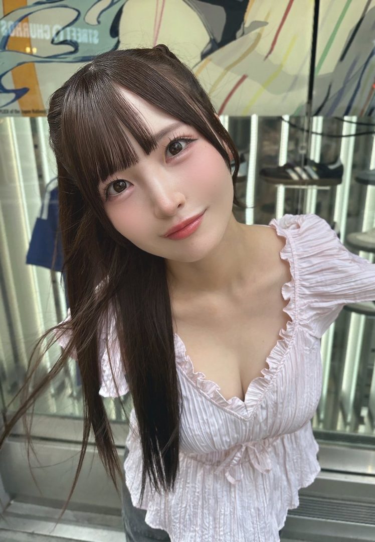 A young woman with long brown hair and bangs, wearing a light pink ruffled blouse, looks directly at the camera with a slight smile.
