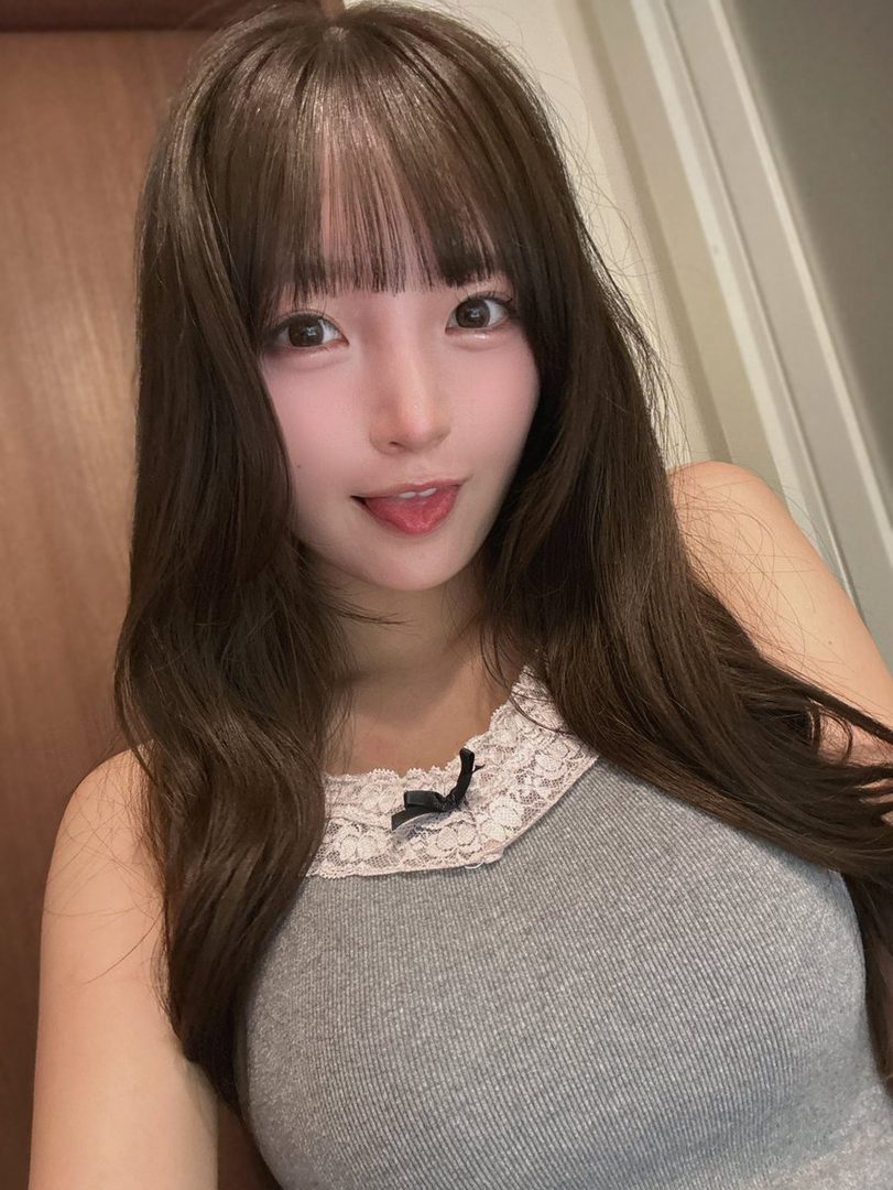 A cute East Asian woman with long brown hair and bangs takes a selfie indoors, sticking out her tongue playfully.