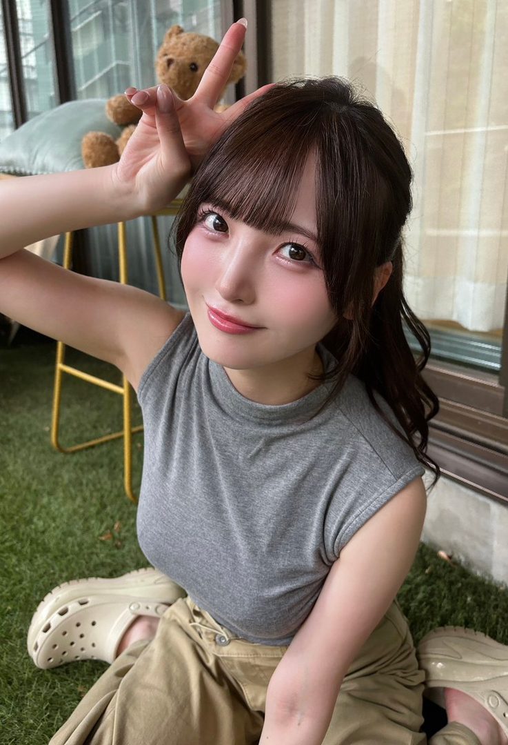 A cute East Asian woman with brown hair and bangs makes a peace sign while sitting indoors on artificial grass.