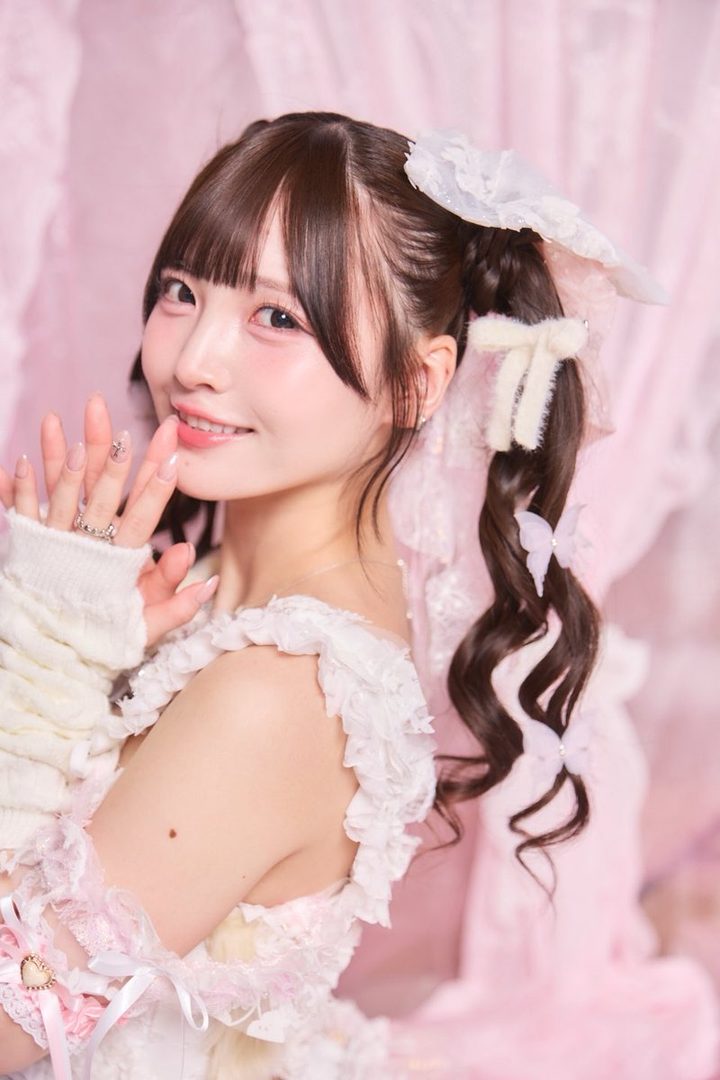 A cute Japanese woman with brown curly twintails and bangs, wearing a white and pink frilly dress, smiles softly at the camera in an indoor setting.