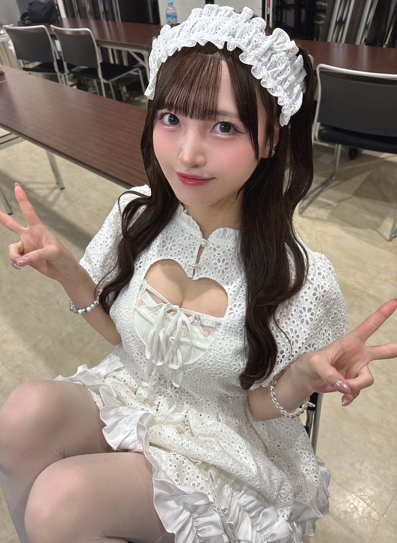 A cute woman with long brown hair and bangs, wearing a white lace dress with a heart-shaped cutout, poses indoors with peace signs.