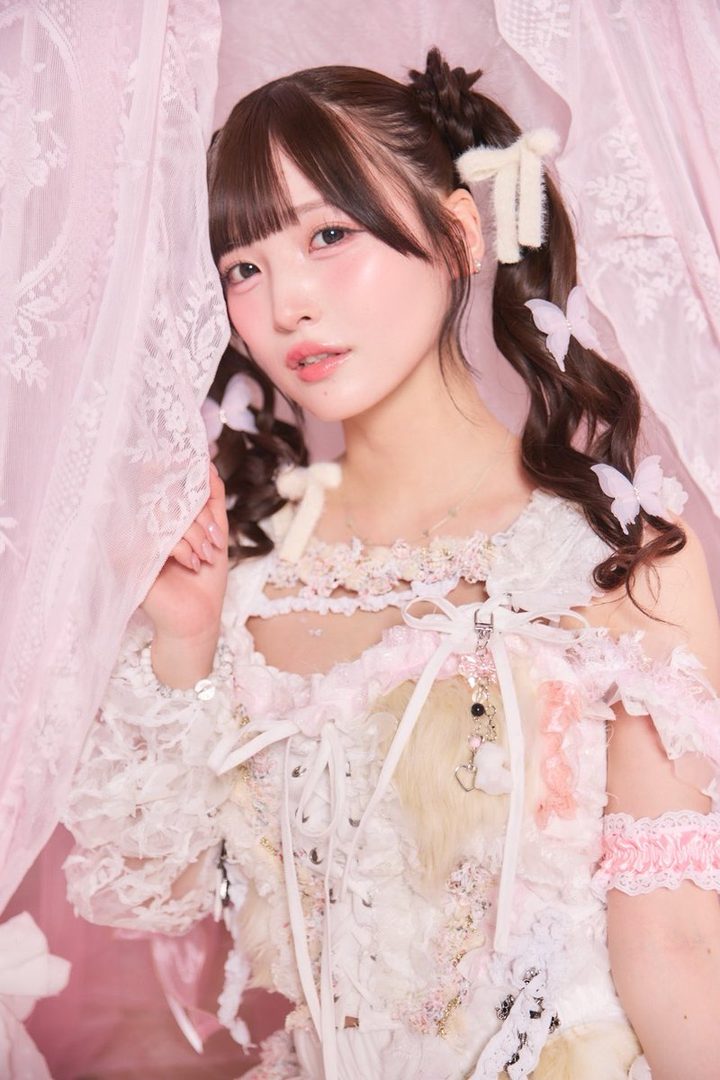A cute Japanese woman with brown twintails and bangs, wearing a pink and white lace dress, peeks from behind a pink sheer curtain.