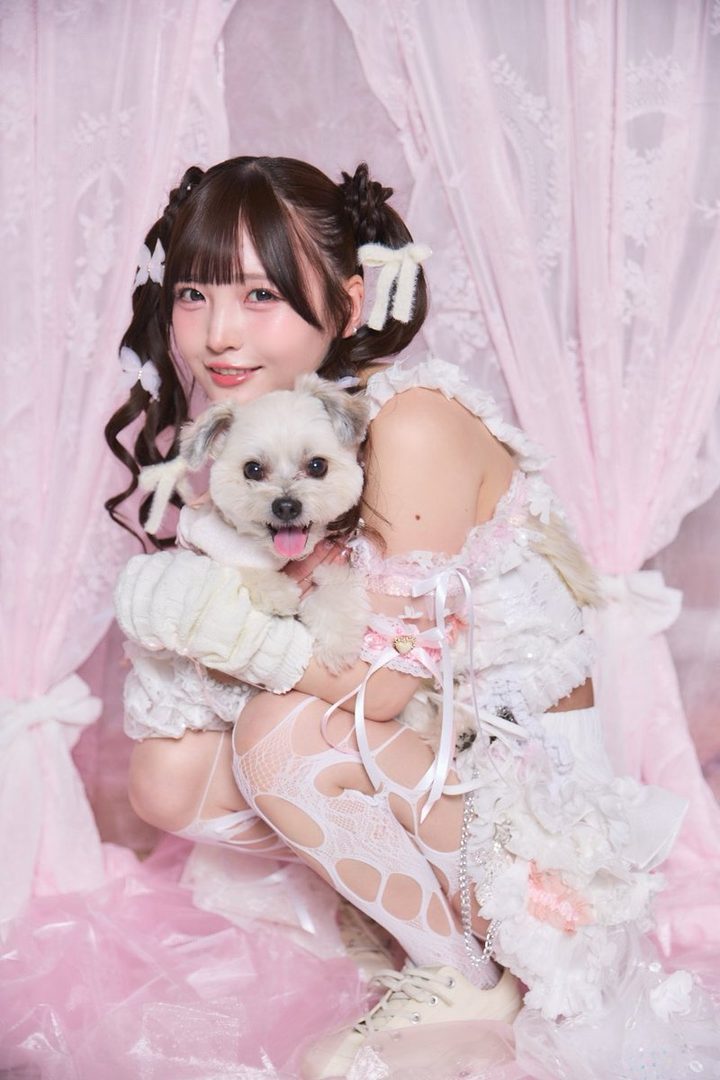 A cute Japanese woman with brown twintails and bangs, wearing a white and pink frilly dress, kneels while holding a small white dog in a pink-themed indoor studio.