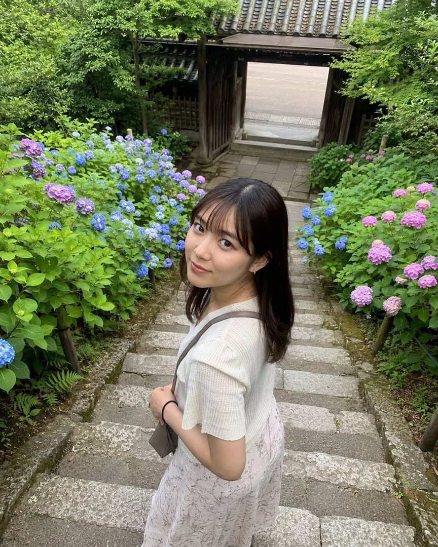 A young woman with long black hair and bangs, wearing a light top and patterned skirt, stands on stone steps surrounded by vibrant blue and purple hydrangeas, looking back at the viewer.