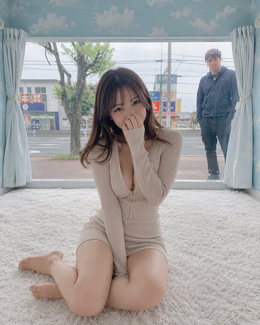 A young woman in a beige knit romper kneels on a white shag rug indoors, covering her mouth with her hand and smiling shyly, with a street scene visible through a large window behind her.