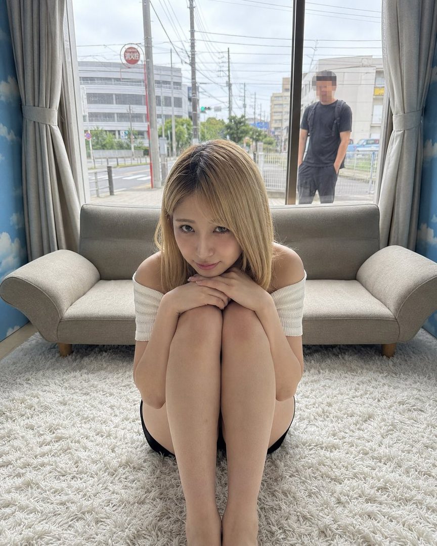 A young woman with blonde hair sits on a white rug indoors, looking at the camera with a gentle expression, with a street view visible through a large window behind her.