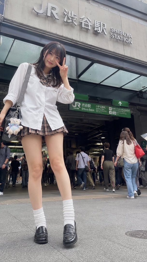 A young woman in a school uniform poses with a peace sign in front of Shibuya Station.