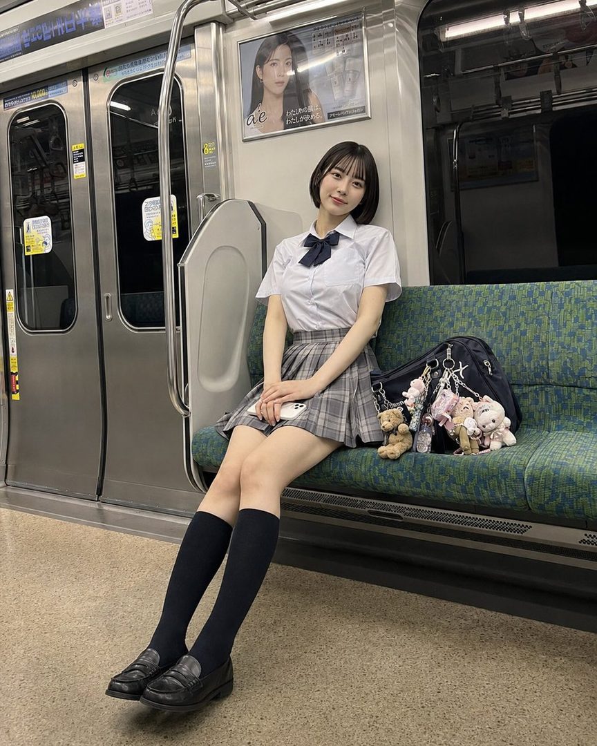A young woman in a Japanese school uniform sits on a train, smiling at the camera.