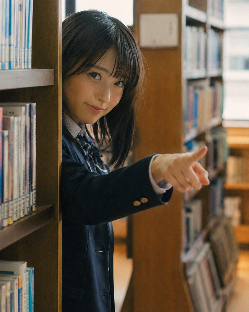 A young woman in a school uniform points towards the viewer from behind a bookshelf in a library.