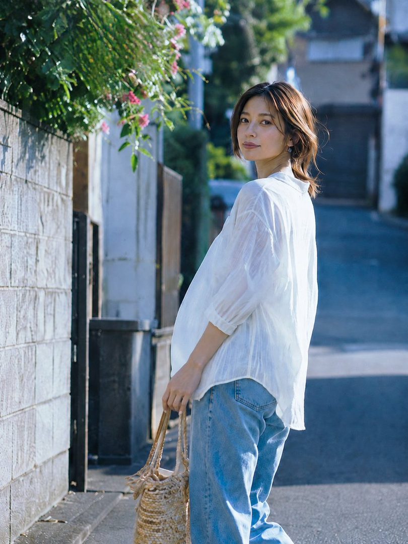 A woman with short brown hair, wearing a white shirt and jeans, walks down a street with a woven bag, looking back at the camera.