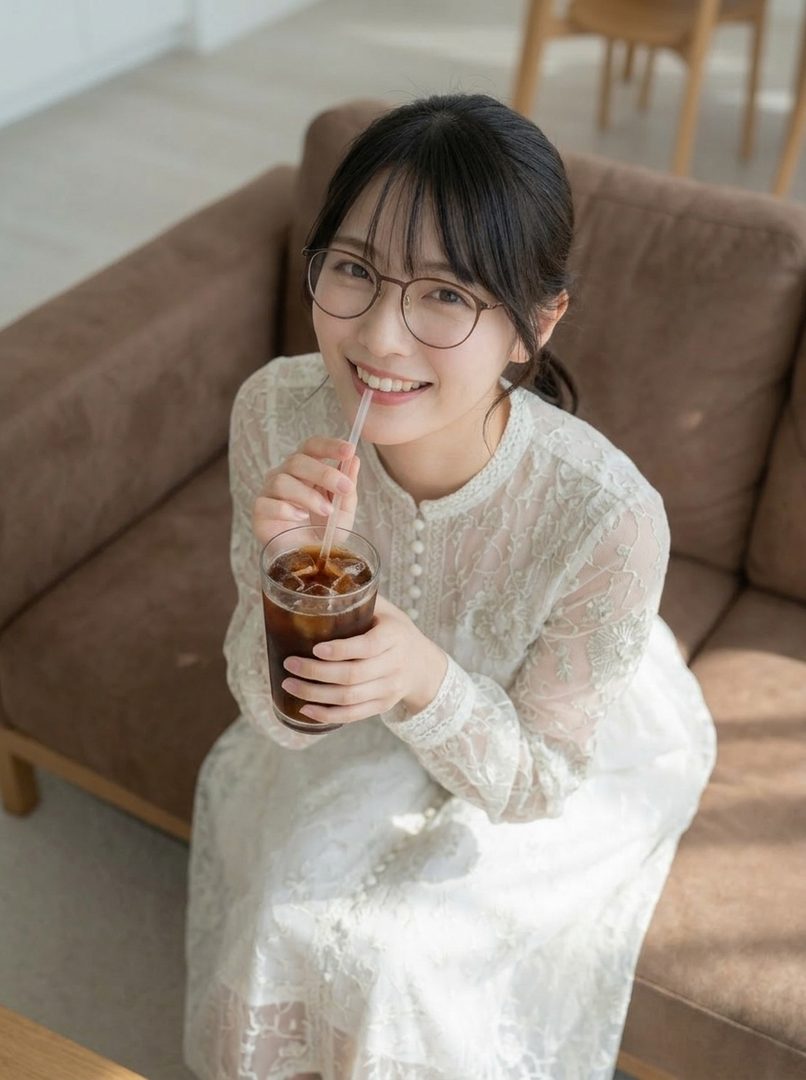 A young woman with black hair and glasses smiles while drinking iced coffee indoors.