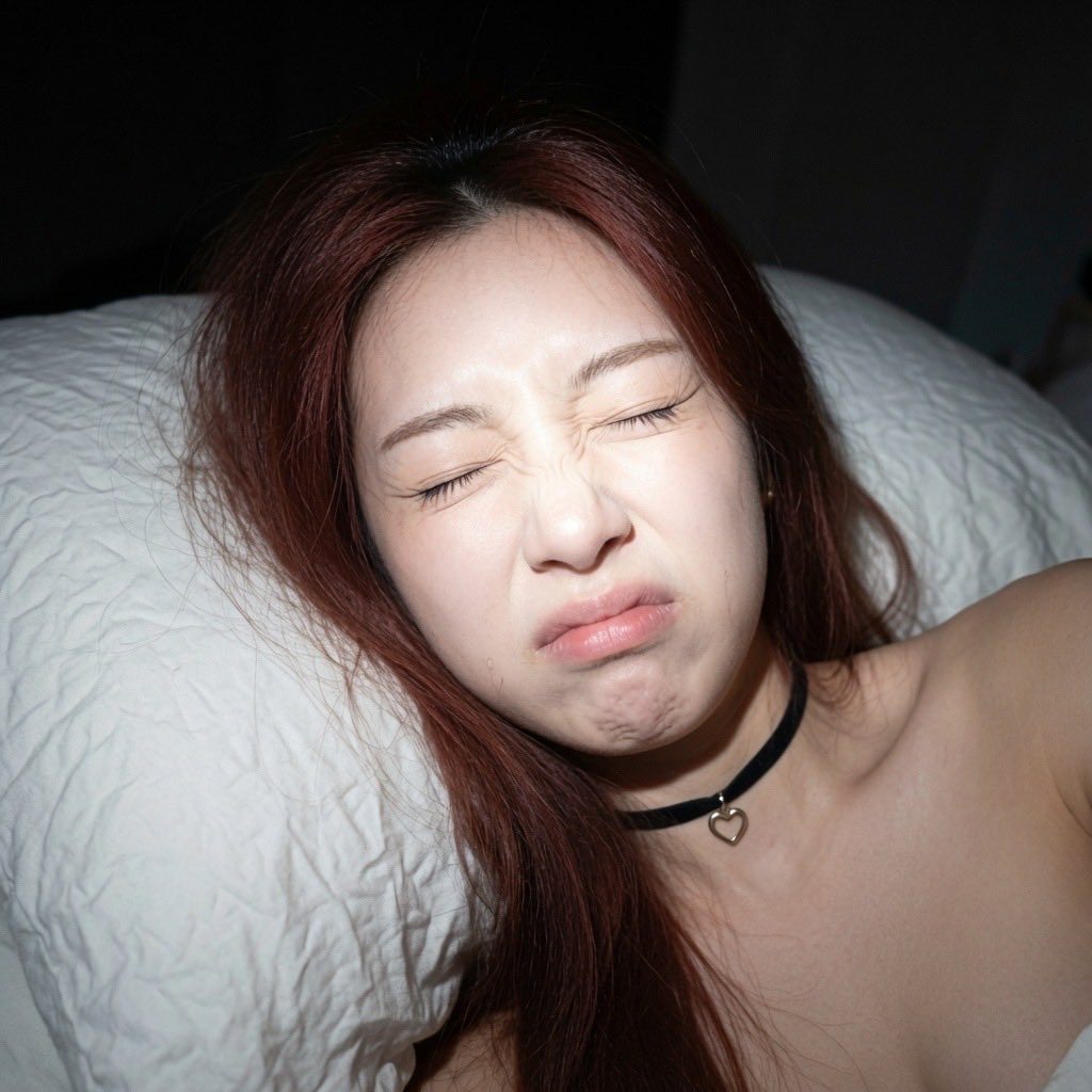 A woman with long red hair lies in bed, making a funny face with her eyes squeezed shut and mouth puckered, wearing a black choker with a heart pendant.