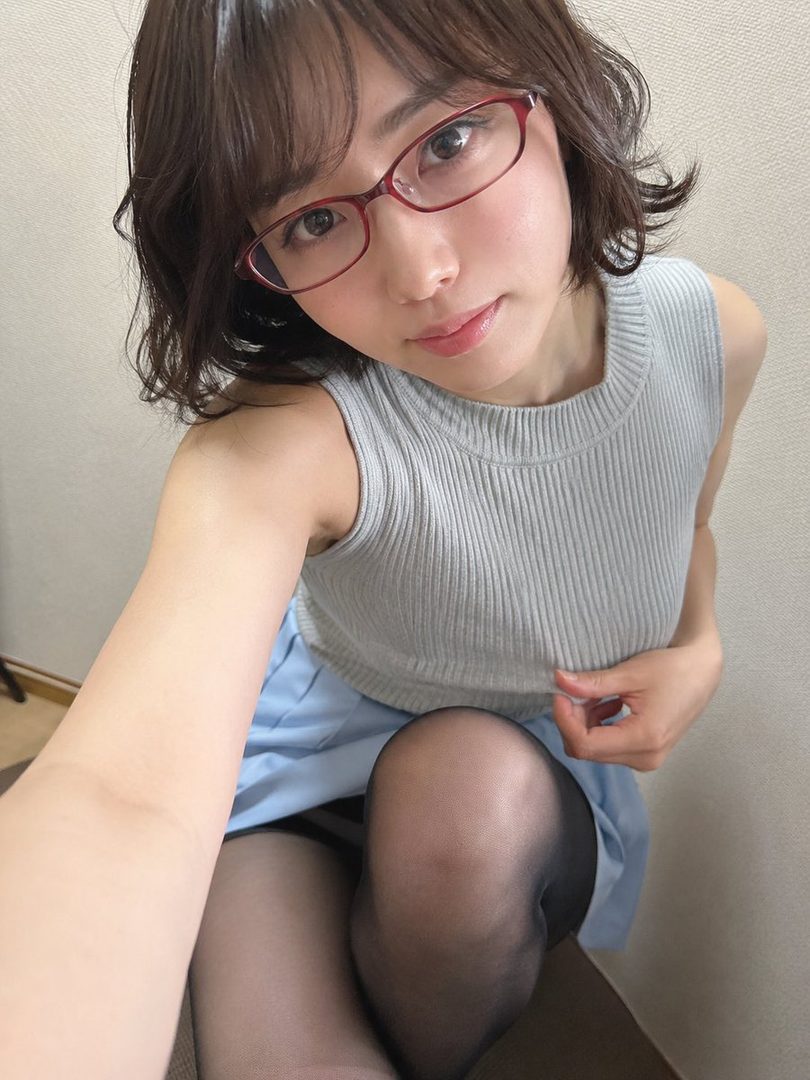 A Japanese woman with short brown hair and red glasses takes a selfie, wearing a light blue knit top and skirt with black stockings.