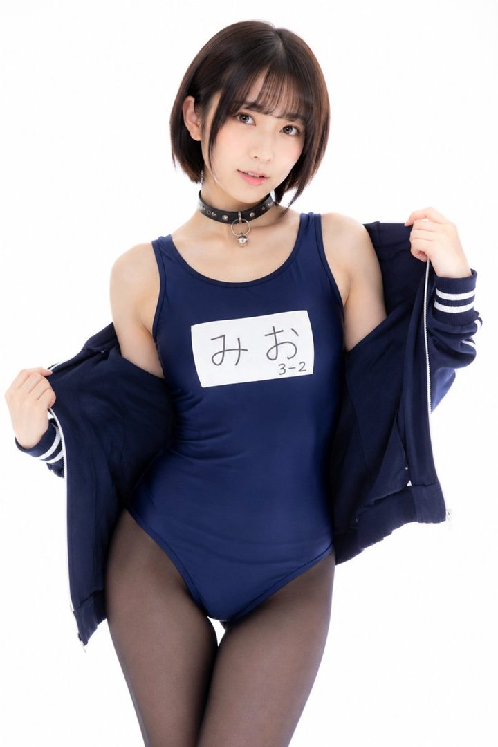 A Japanese woman with short black hair and bangs poses in a navy blue one-piece swimsuit and black tights, with a matching jacket draped over her shoulders, against a white studio background.