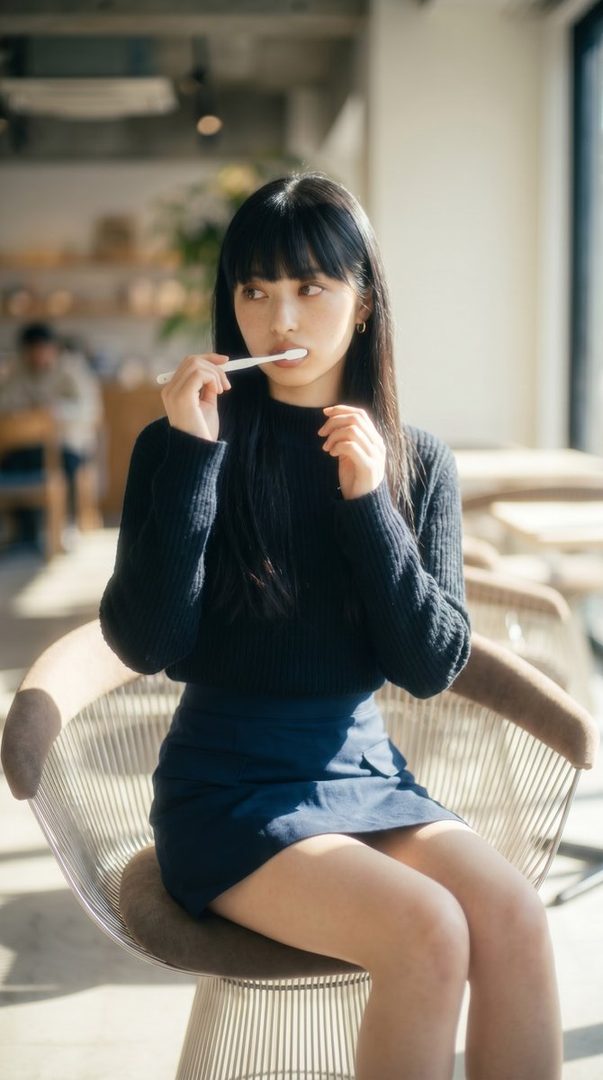 A young woman with black hair and bangs, wearing a black sweater and dark skirt, sits indoors and holds a toothbrush to her mouth.