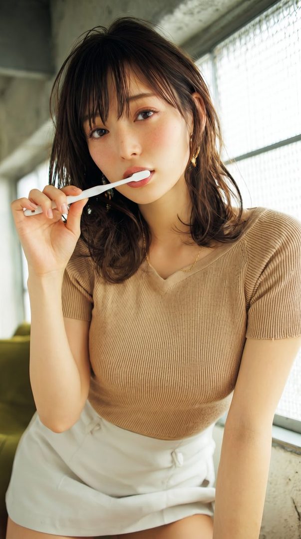 A young woman with brown hair and bangs, wearing a beige top and white shorts, holds a toothbrush to her mouth indoors.