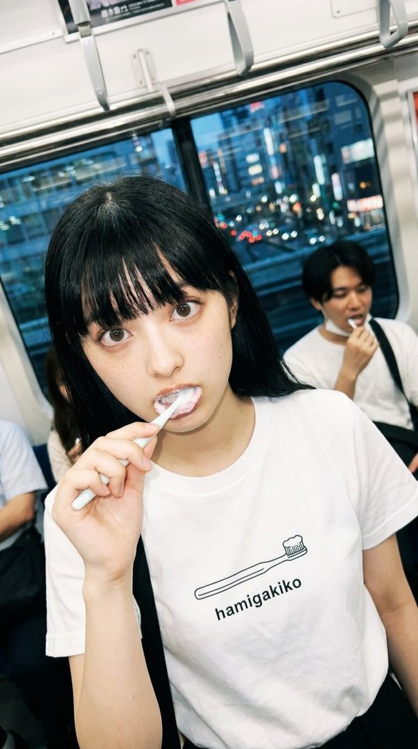 A young woman with black hair and bangs brushes her teeth on a train, wearing a white t-shirt with a toothbrush graphic.