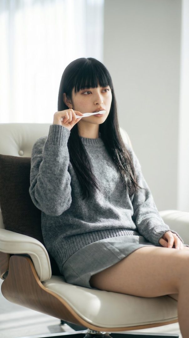 A young woman with long black hair and bangs, wearing a grey sweater, sits in a chair and brushes her teeth indoors.