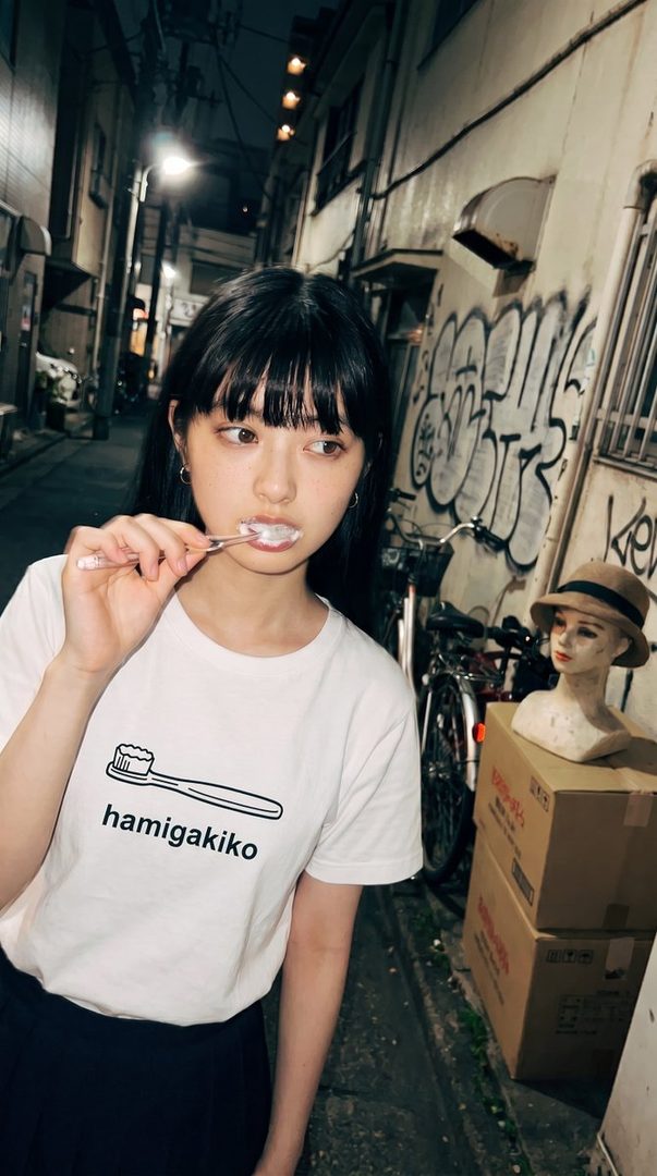 A young woman with black hair and bangs brushes her teeth on a dark street at night, wearing a white t-shirt with a toothbrush graphic.