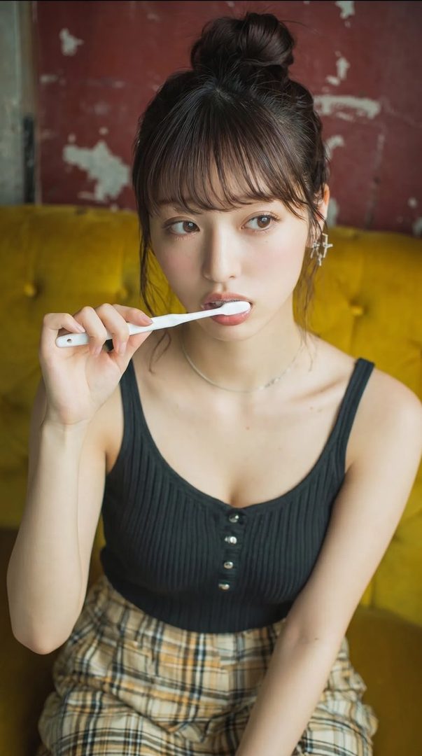A young woman with brown hair in a bun and bangs, wearing a black tank top and plaid skirt, holds a toothbrush to her mouth while looking to the side.