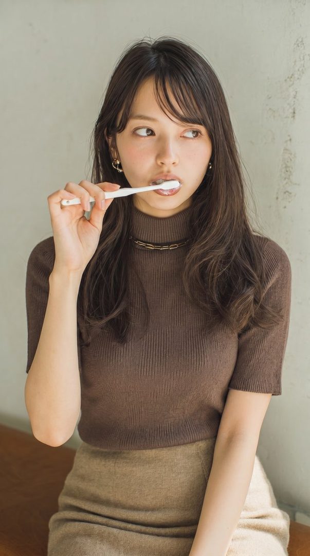 A young woman with long brown hair is brushing her teeth while looking to the side, wearing a brown knit top and a beige skirt.