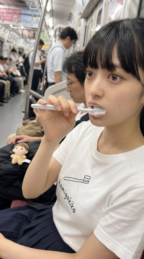 A young woman with black hair and bangs is brushing her teeth on a train, wearing a white t-shirt with a toothbrush graphic and a dark pleated skirt.