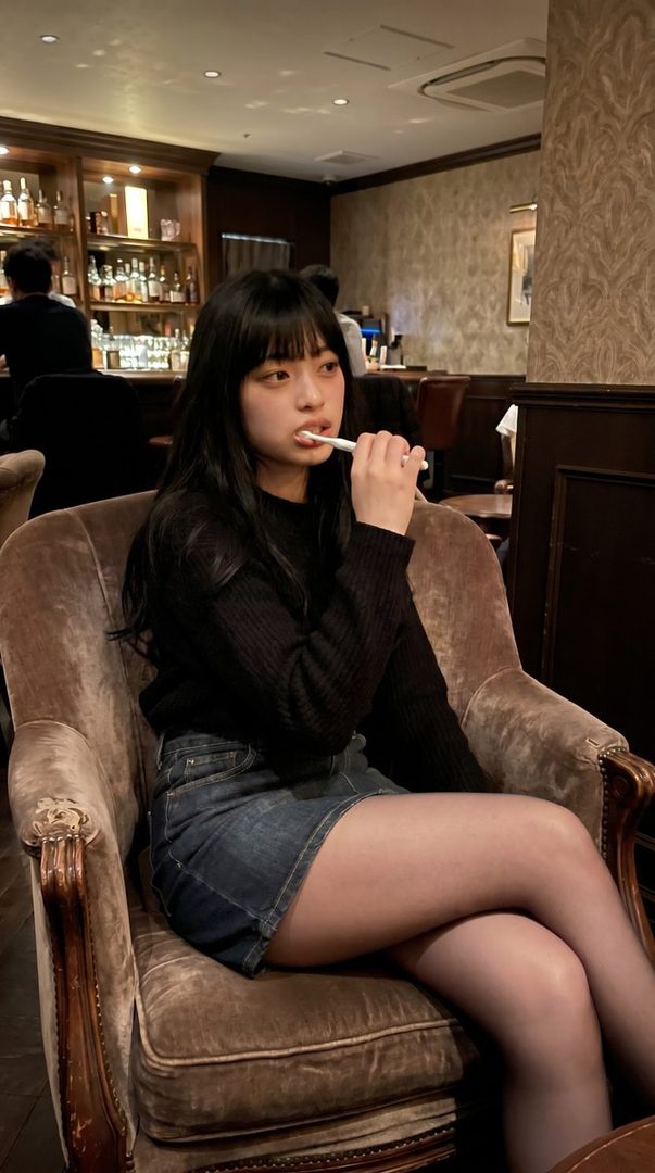 A young woman with long black hair and bangs, wearing a black knit top and a denim skirt, sits in a plush armchair indoors, holding a toothbrush to her mouth.