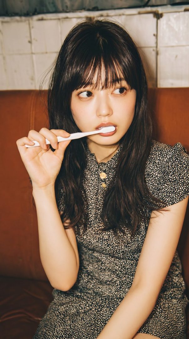 A young woman with dark hair and bangs is brushing her teeth while looking to the side, wearing a patterned dress indoors.
