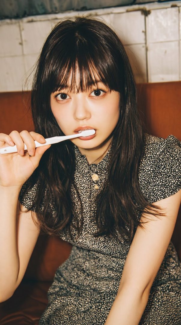 A young woman with long dark hair and bangs, wearing a patterned dress, holds a toothbrush to her mouth indoors.