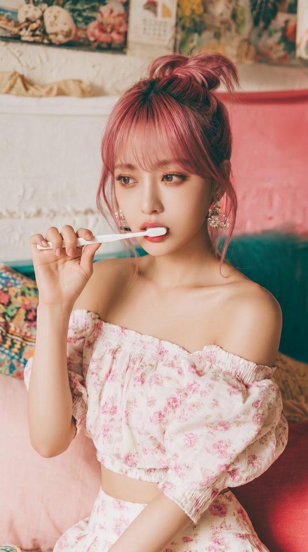 A young woman with pink hair and a floral off-the-shoulder top holds a toothbrush to her mouth.