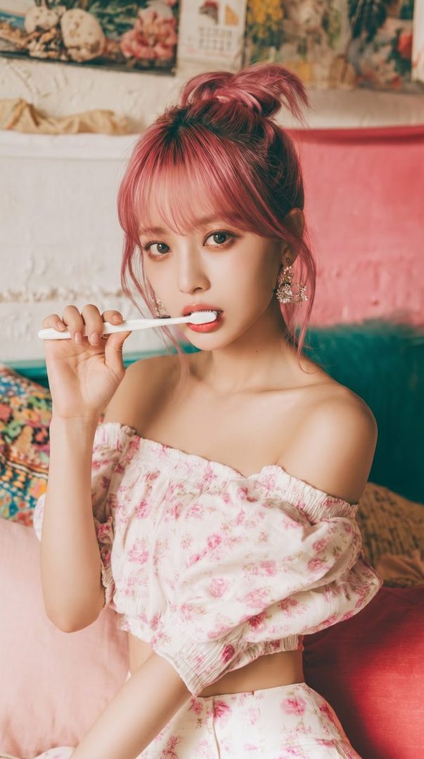 A young woman with pink hair and a floral off-the-shoulder top holds a toothbrush to her mouth, looking at the camera.