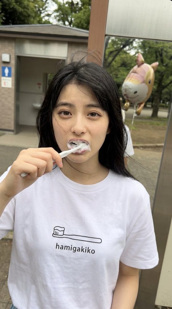 A young woman with black hair brushes her teeth outdoors, wearing a white t-shirt with a toothbrush graphic.