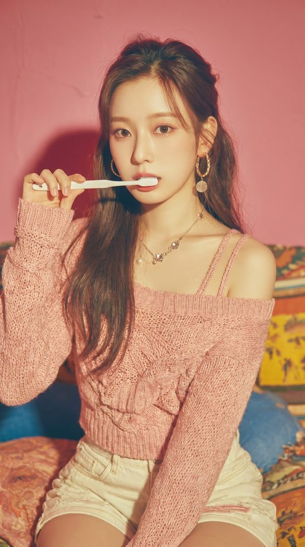 A young woman with long brown hair and a pink knitted top holds a toothbrush to her mouth, looking directly at the camera.