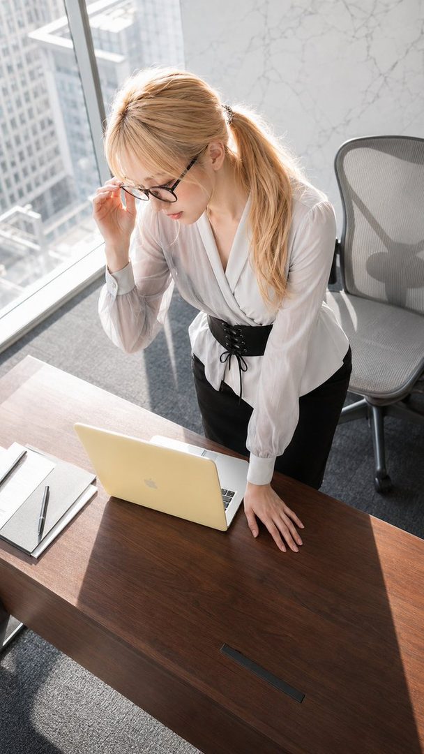 A blonde woman in a white blouse and black skirt is working on a laptop in a modern office with a city view.