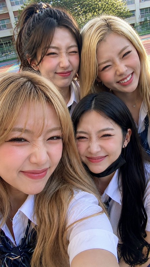 Four young women in school uniforms take a playful selfie outdoors, smiling and winking at the camera.