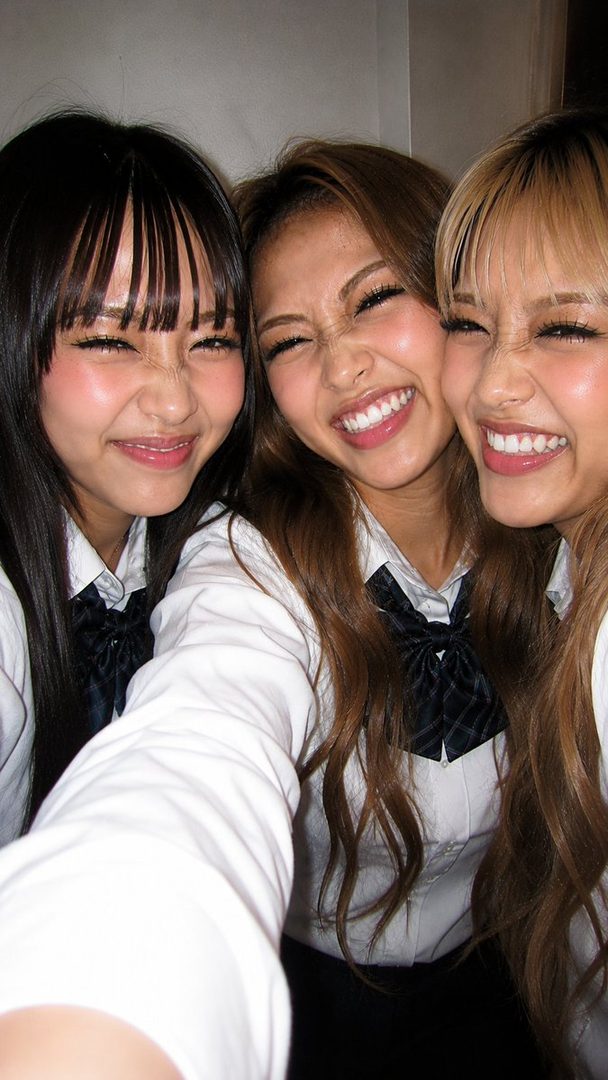 Three young women in school uniforms take a playful selfie, smiling widely with crinkled eyes.