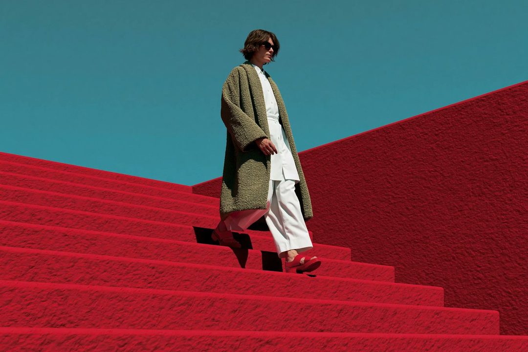 A person in a long green coat and white outfit walks down bright red stairs under a clear blue sky.
