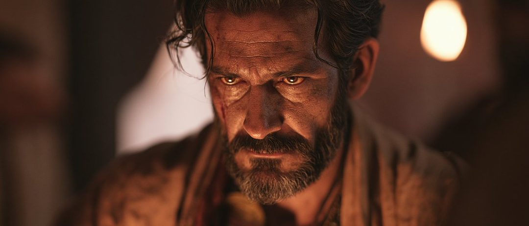 A close-up portrait of a rugged man with a beard and intense eyes, illuminated by warm, dramatic lighting.