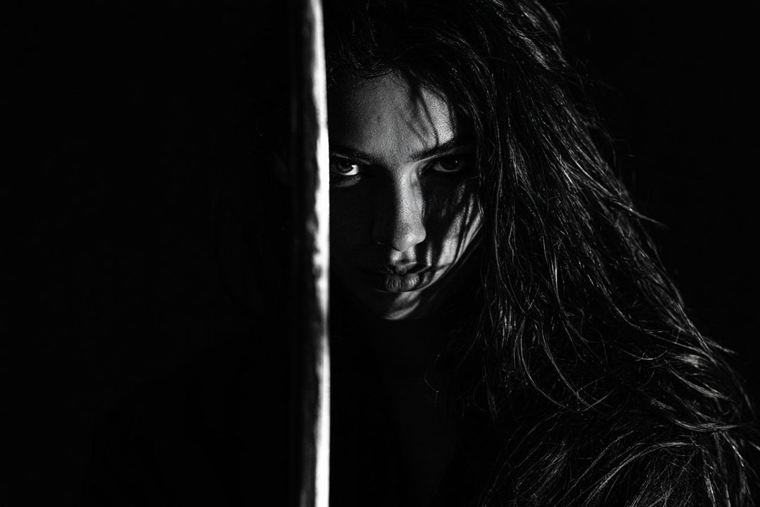 A dramatic black and white portrait of a woman with long dark hair, partially obscured by shadow and a vertical object, looking intensely at the viewer.