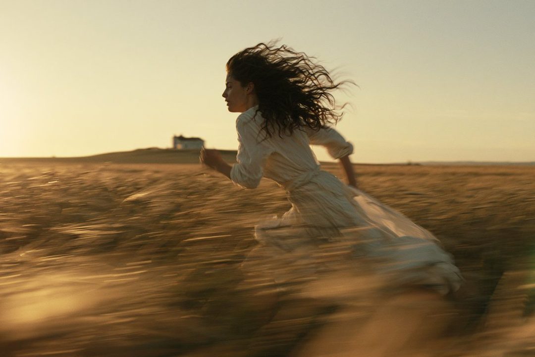 A woman with long, curly brown hair runs through a golden field at sunset, wearing a white dress.