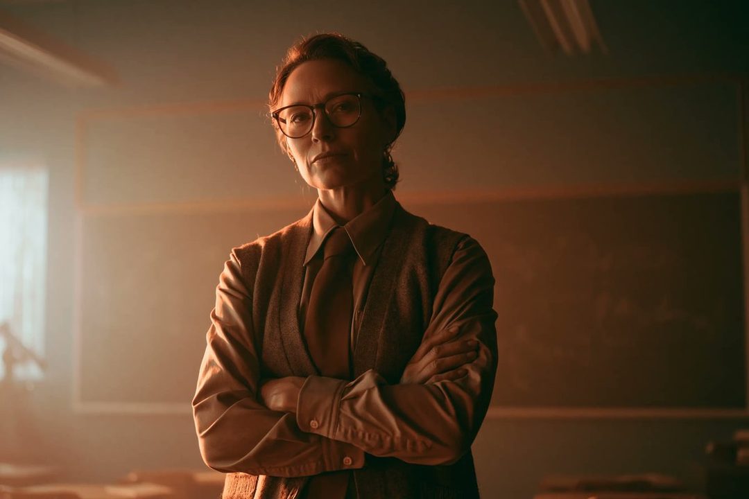 A photorealistic portrait of a woman with glasses and a tie, standing in a classroom with her arms crossed, evoking a 1970s aesthetic.