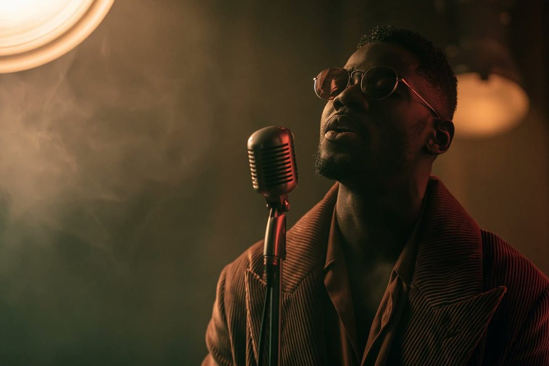 A man wearing sunglasses and a striped jacket sings into a vintage microphone in a dimly lit, smoky room.