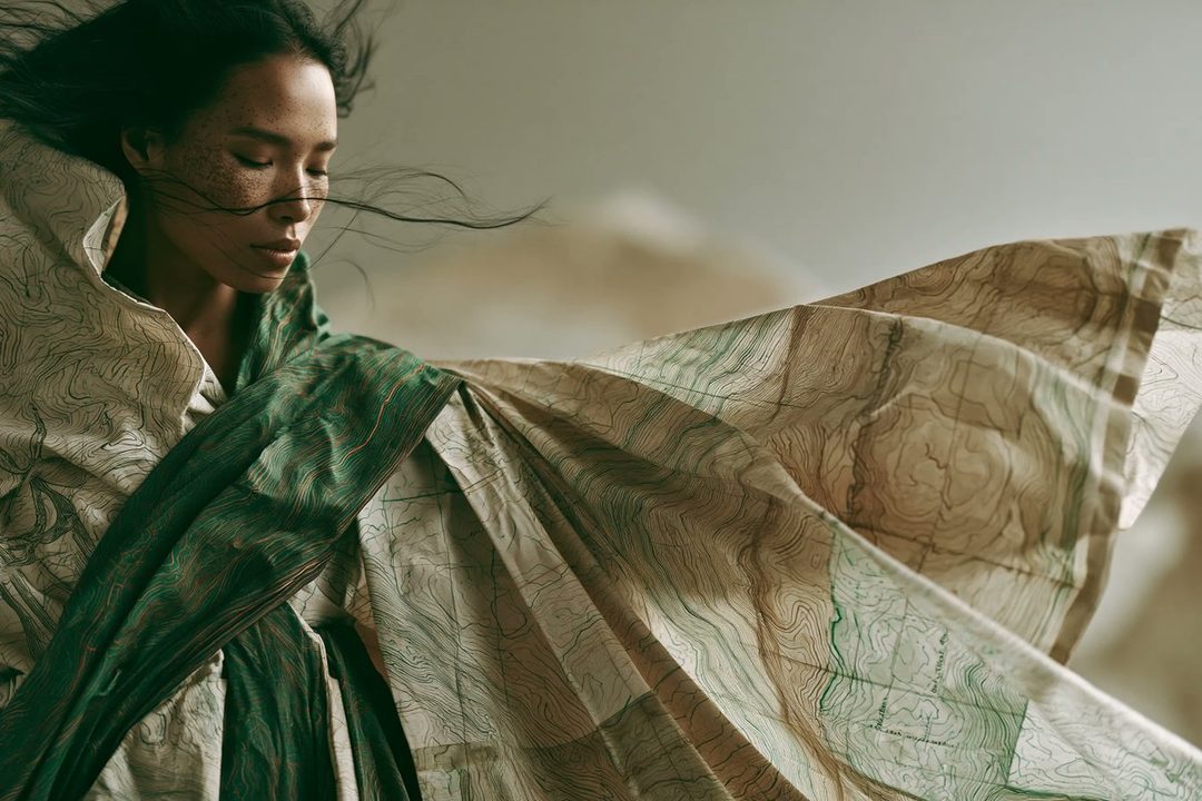 A woman with dark hair and freckles on her face, wearing an avant-garde garment made from flowing topographic maps, with wind-blown hair.