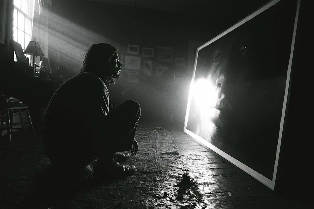 A person with long hair crouches in a dark room, looking at a large, brightly lit portrait of a face, with rays of light streaming in from a window.