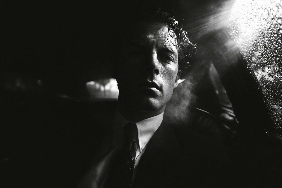 A dramatic black and white portrait of a man in a suit, with half his face in shadow and light reflecting off a wet window.